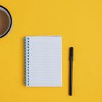 notepad and black pen with cup of coffee with cream on yellow background