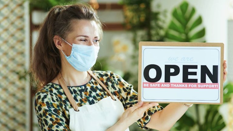 woman wearing a mask and holding an open sign to open up her business for the day